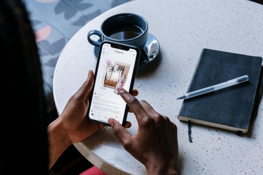 Hands holding smartphone at a cafe table with coffee and a notebook, focusing on social media browsing.